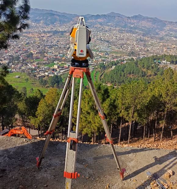 A surveying total station on a tripod stands atop a hill, with a clear view of a densely populated valley and distant mountains